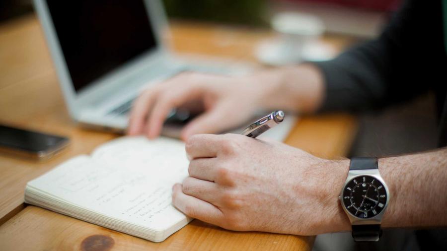 Person writing notes next to a laptop